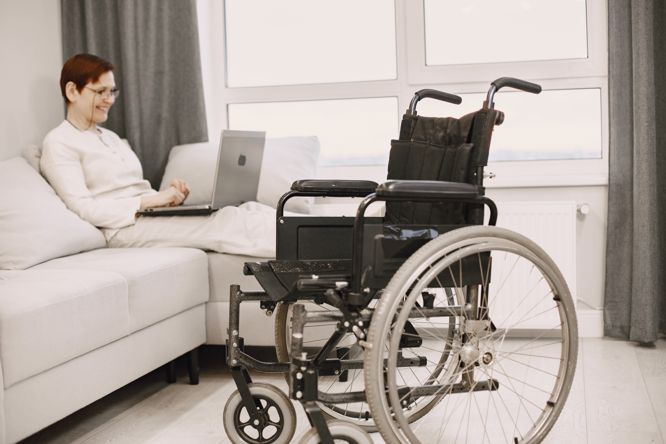Smiling Woman Sitting on a Couch and Using Laptop and a Wheelchair Standing Next to the Couch 
